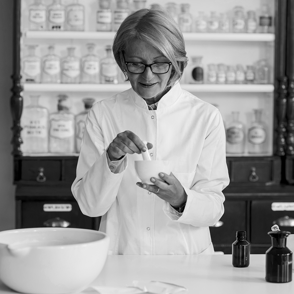 Person in a white coat preparing ingredients in a bowl with shelves of jars in the background